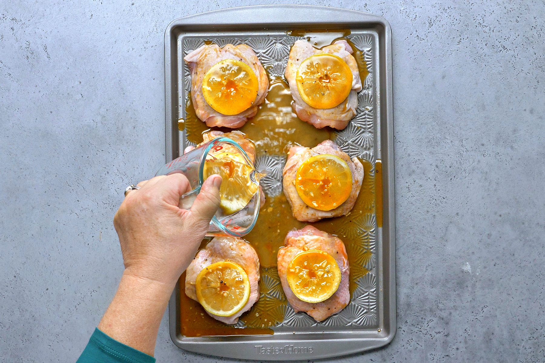 A person's hand pouring marinade from a measuring cup over six raw chicken thighs topped with lemon slices on a baking sheet. The sheet is on a gray countertop.