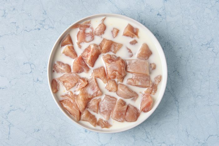 A bowl filled with chunks of raw chicken marinating in buttermilk on a light blue marbled surface.