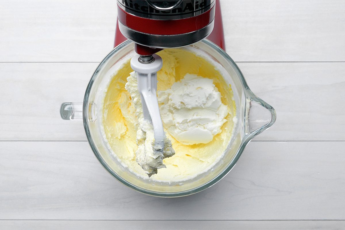 A red stand mixer with a glass bowl is mixing cream or batter. The mixture inside is light and fluffy, showing texture from the mixing process. The setup is on a white wooden surface.