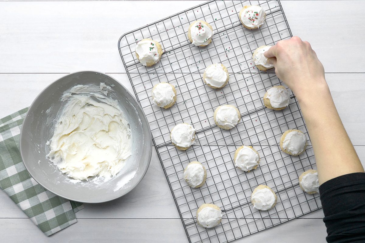 A person is decorating cookies with white icing on a cooling rack. A bowl filled with icing and a green checked cloth are placed beside the rack. The background is a white wooden surface.