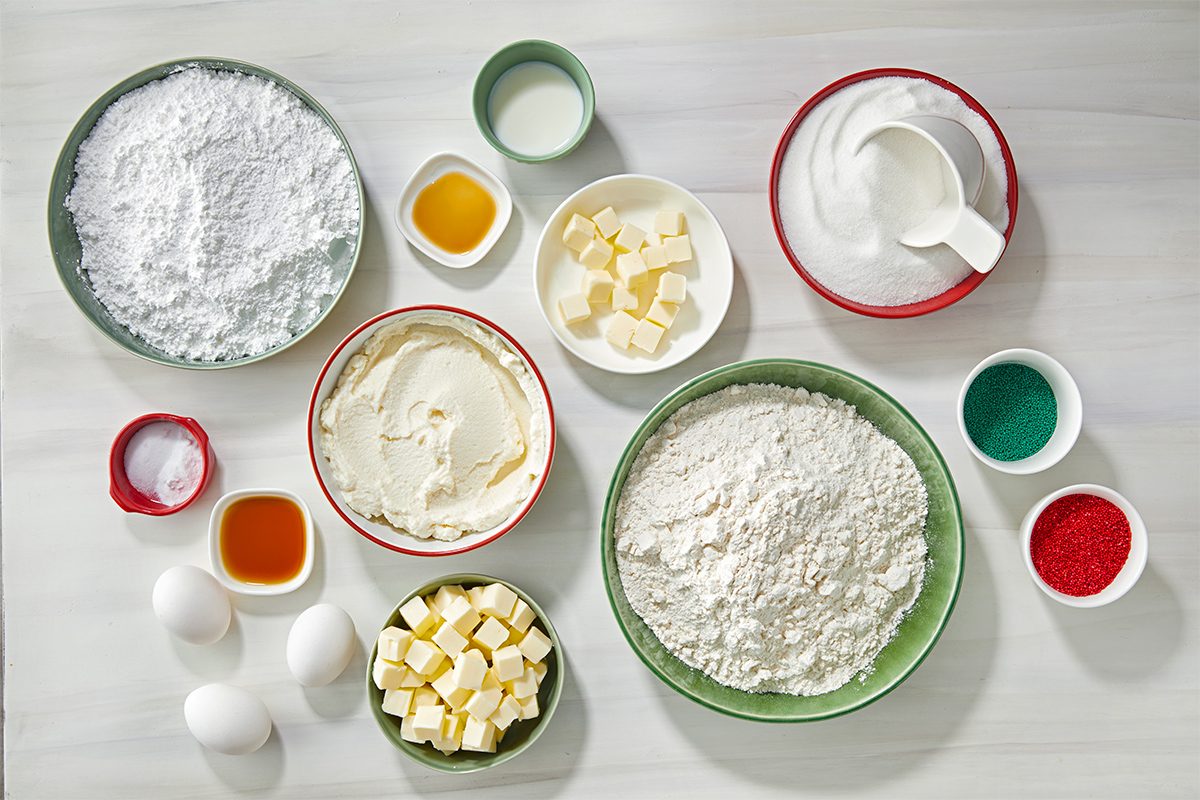 Top-down view of baking ingredients arranged on a white surface, including flour, sugar, butter cubes, eggs, vanilla extract, baking soda, milk, and bowls of red and green food coloring.