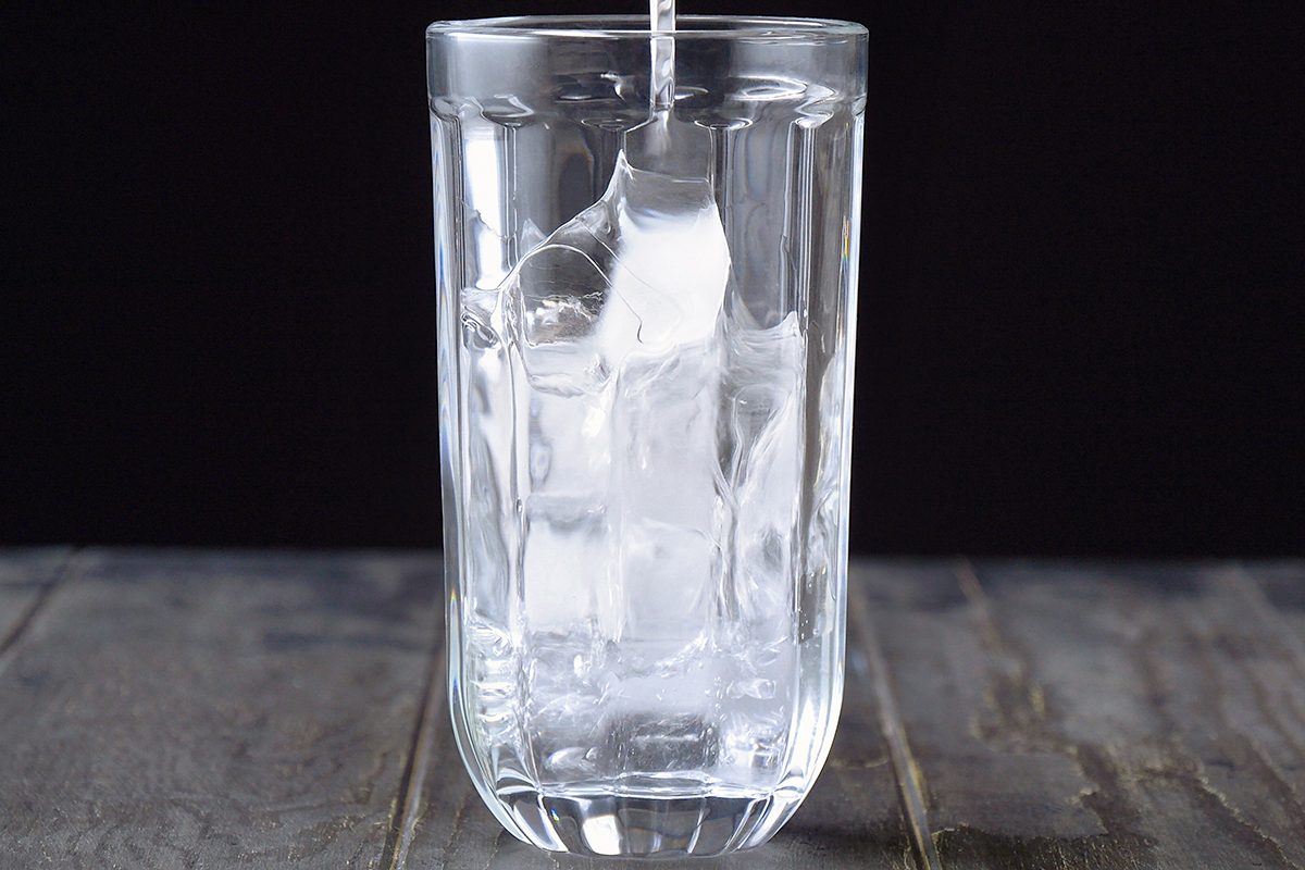A clear glass filled with ice cubes is being filled with water against a dark background. The glass sits on a wooden surface, and the water is creating ripples as it pours in.