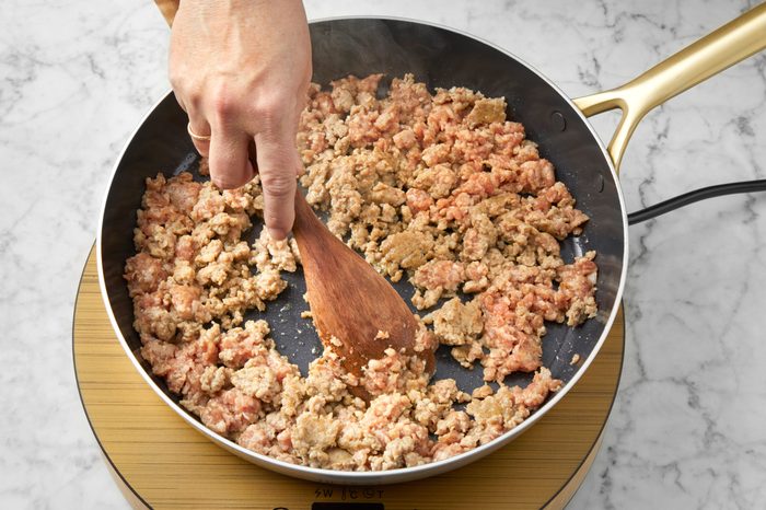 A hand stirs ground meat in a non-stick frying pan using a wooden spatula. The pan is placed on a circular hot plate over a marble countertop. The meat is partially cooked, showing a mix of brown and pink tones.