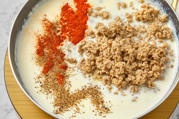 A skillet filled with a creamy white sauce, topped with ground meat, red paprika, and brown spices. The ingredients are partially mixed with a visible swirl pattern.