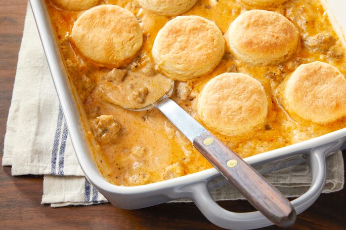 A casserole dish filled with golden-brown biscuits on top of a creamy, sausage gravy mixture. A serving spoon rests in the dish, which is on a striped cloth over a wooden table.