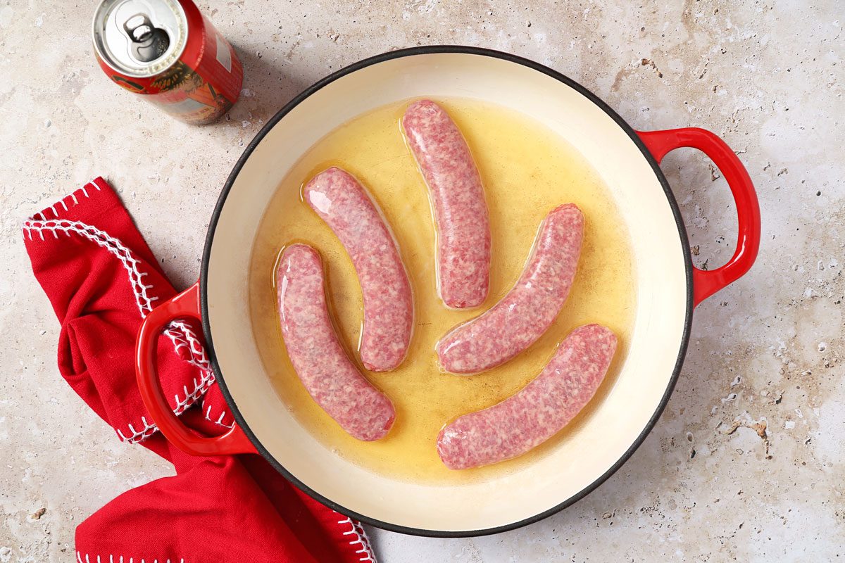 overhead shot of a red enameled cast iron skillet with a red handle; the skillet contains five raw sausages in a bath of oil; the sausages are arranged in a loose circle with the ends of the sausages pointing inwards; the skillet rests on a light brown speckled countertop with a red cloth napkin with white crocheted edges in the foreground