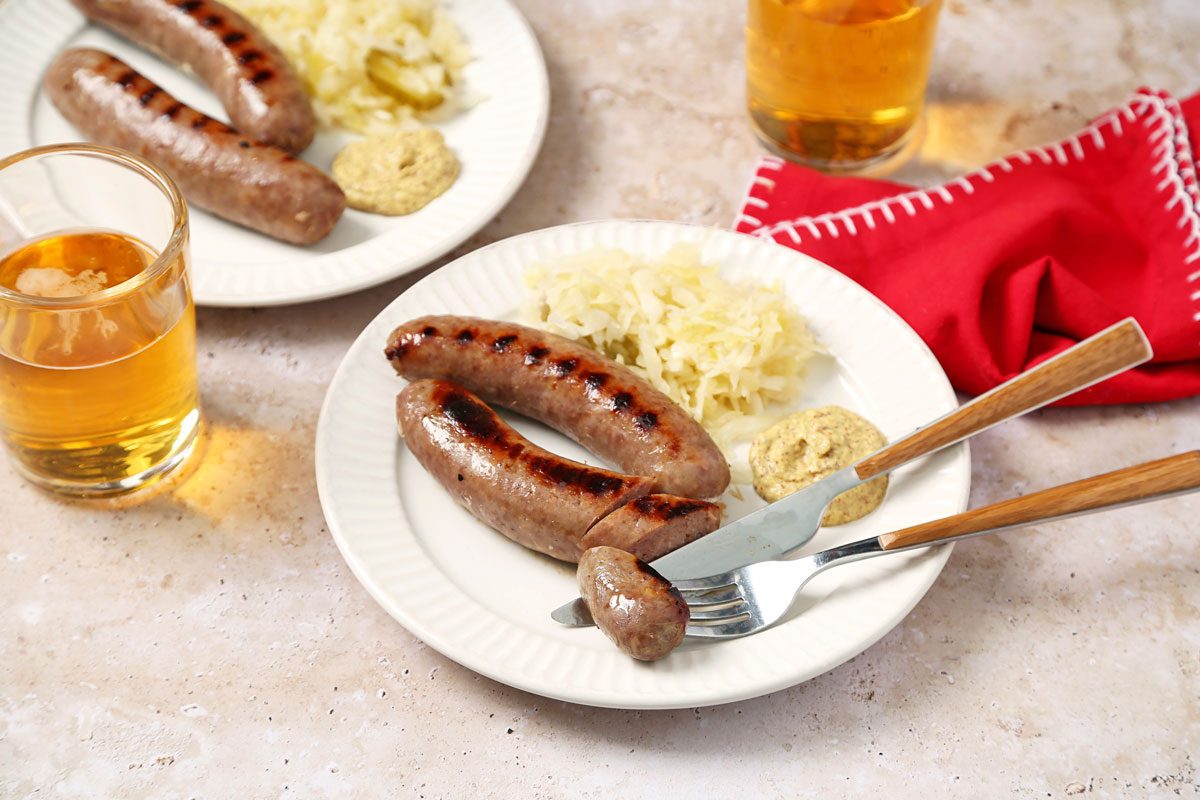 3/4th shot of a plate of food on a light-colored countertop; two sausages, a mound of sauerkraut, and a dollop of mustard are on the plate; a glass of beer is in the foreground; a red napkin is in the background