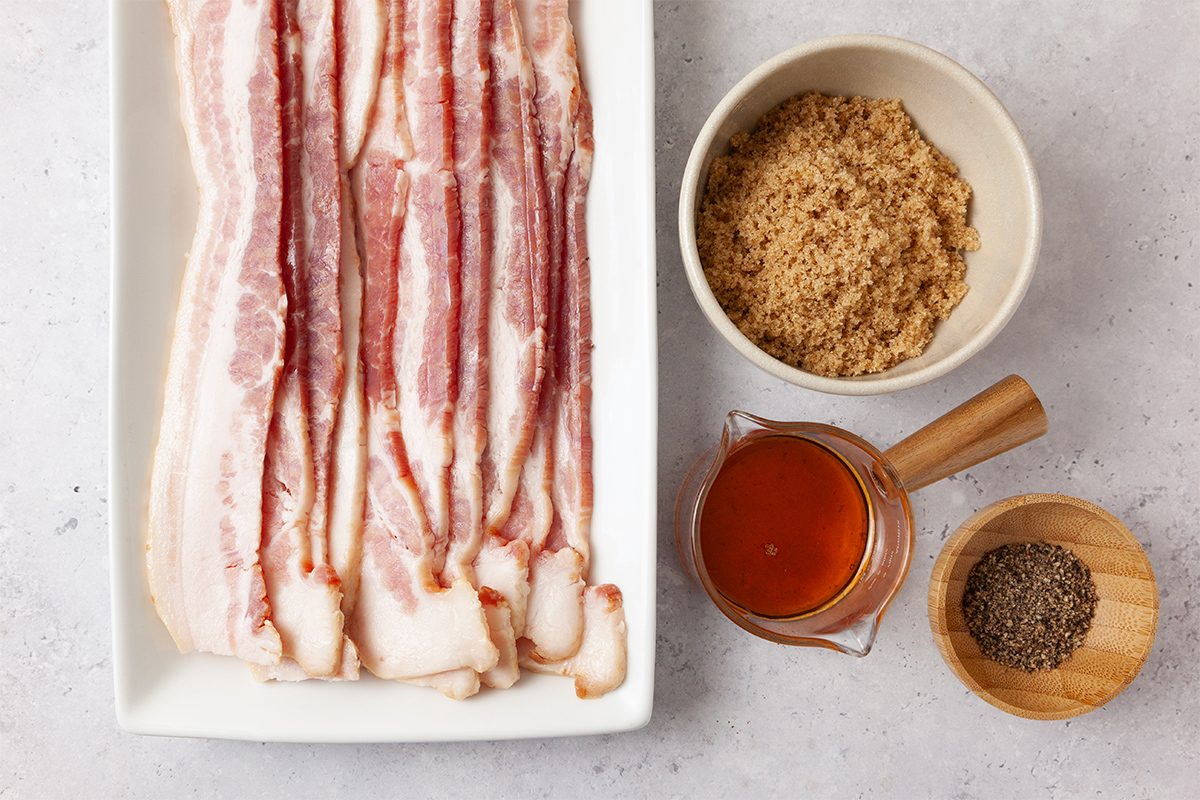 Rectangular plate with raw bacon strips next to a bowl of brown sugar, a small pitcher of hot sauce, and a wooden bowl of black pepper on a light gray surface.