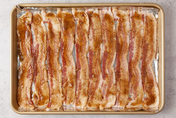 Strips of bacon are arranged in rows on a baking sheet lined with foil, seasoned with brown sugar and spices, ready for cooking.