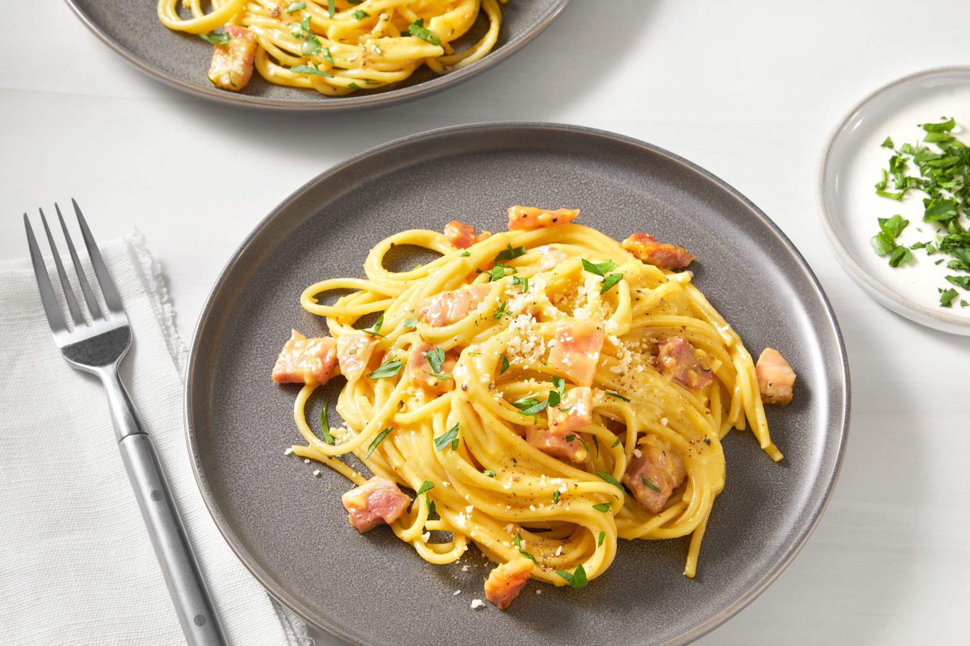 Two plates of carbonara spaghetti topped with crispy bacon and fresh herbs