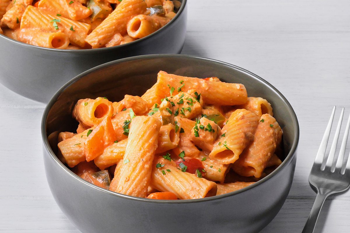 Two bowls of creamy rigatoni pasta garnished with chopped parsley. The pasta is mixed with a rich, orange-hued sauce and visible pieces of vegetables. A fork is placed next to one of the bowls on a light wooden surface.