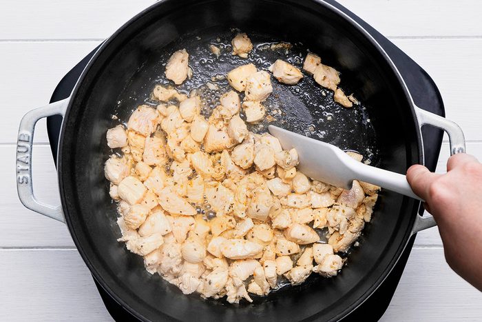 A person stirring diced chicken cooking in a black pot with a white spatula. The chicken is lightly seasoned and simmering in oil, with steam rising. The pot handle reads "Staub." The background is a white surface.