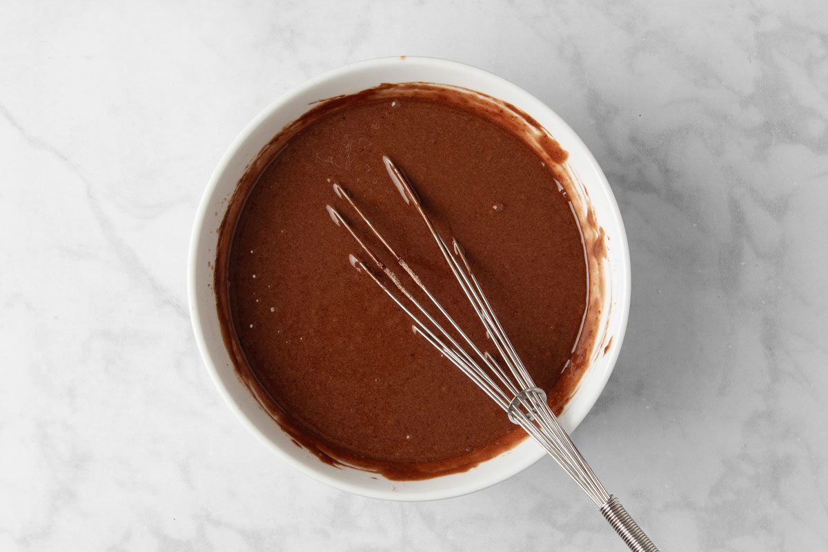 overhead shot of a white bowl containing a thick chocolate batter, a whisk lays beside the bowl; the bowl is sitting on a white marble surface with subtle gray veining
