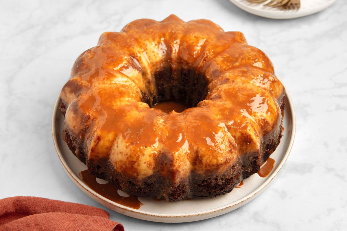 closeup shot of a baked bundt cake with a caramel glaze; the cake is sitting on a white plate and is surrounded by a white marble countertop; a small piece of orange fabric can be seen in the lower left corner of the image