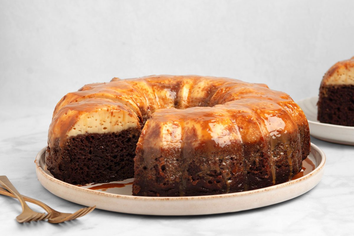 horizontal shot of a Chocoflan, on a white plate with two gold forks; a second slice of Chocoflan sits on another white plate in the background