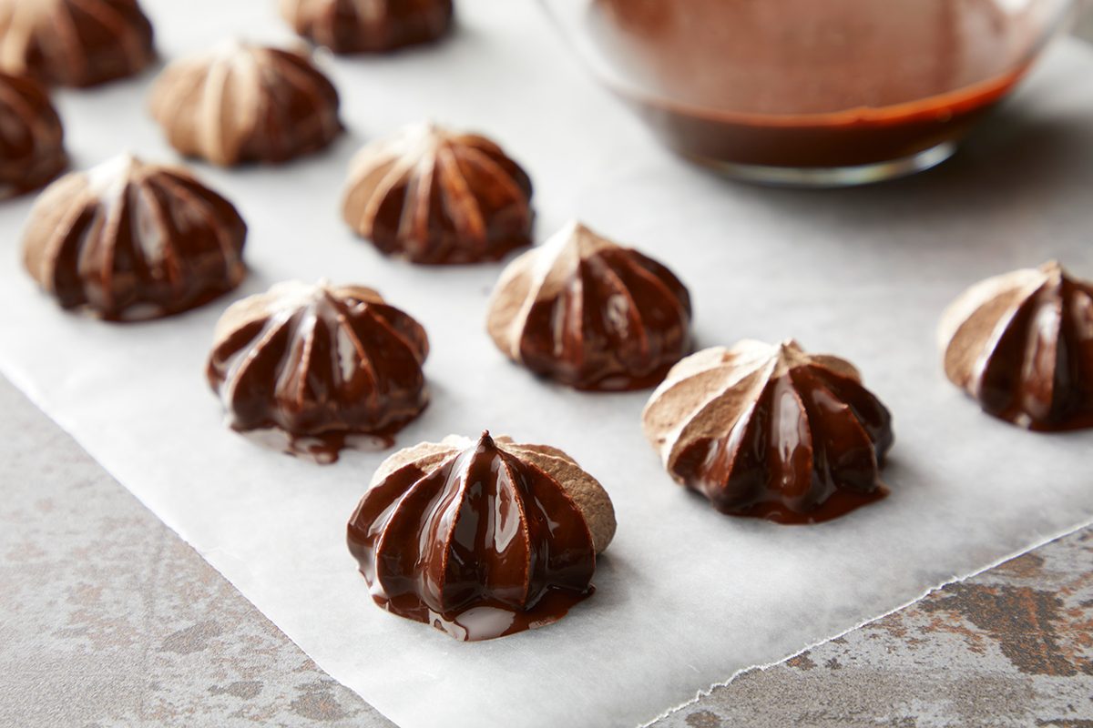 Chocolate meringue cookies on parchment paper, each dipped in melted chocolate. A bowl of chocolate sauce is visible in the background. The cookies have a swirled texture and are arranged in neat rows.