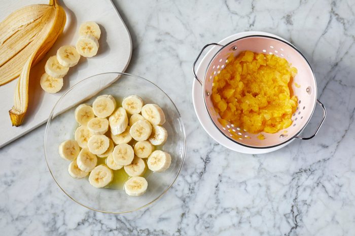 Sliced bananas on a plate, a peeled banana on a cutting board, and crushed pineapple in a colander on a marble countertop.