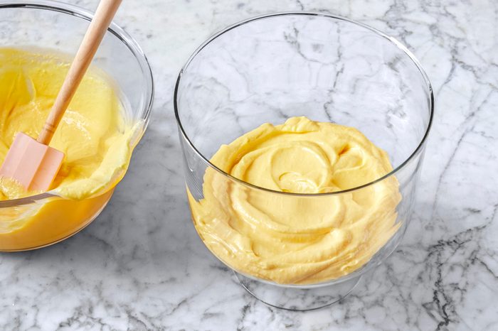 A glass bowl on a marble surface containing whipped yellow cream swirled smoothly. Next to it, another bowl with more whipped cream and a pink spatula.