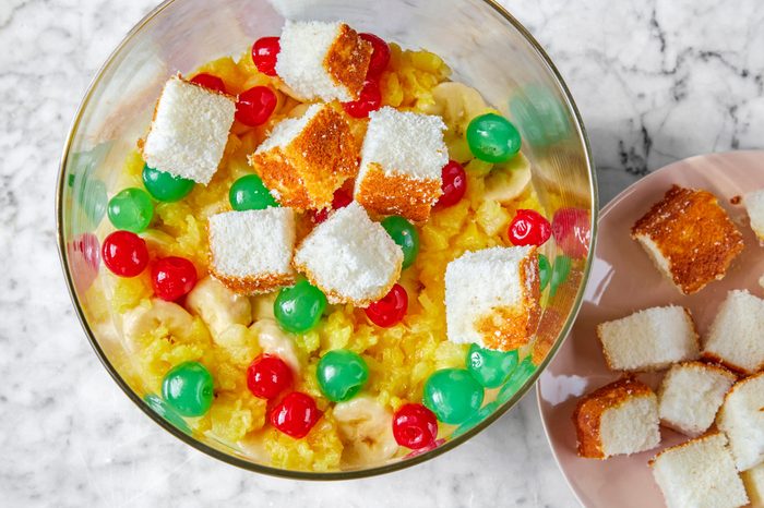 A glass bowl filled with a colorful dessert of cubed cake pieces topped with shredded coconut, surrounded by red and green maraschino cherries, and a mixture of pineapple and banana slices. A few extra cake cubes are on a plate nearby.