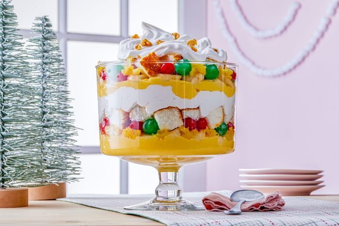 A festive trifle in a glass bowl featuring layers of whipped cream, cake, colorful jelly beans, and yellow custard. It sits on a table with a small spoon and pink plates nearby. A decorative tree and window are in the background.