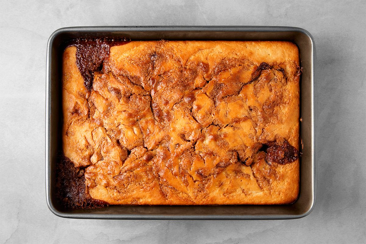 A rectangular baking dish containing a golden-brown cake with a swirled cinnamon pattern on top, viewed from above on a gray surface.