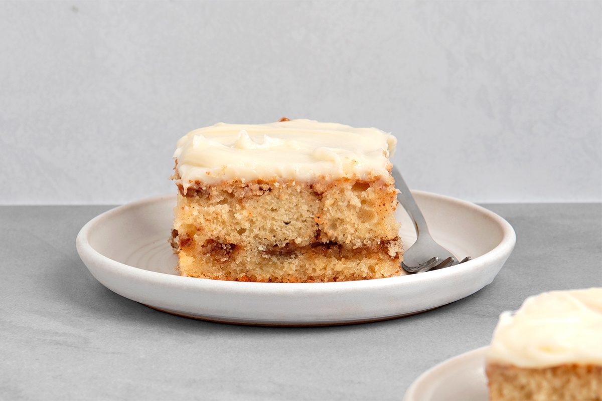 A slice of cinnamon roll cake with cream cheese frosting sits on a white plate. A fork rests on the plate, suggesting the cake has been partially eaten. The background is a light gray surface, creating a simple and clean presentation.