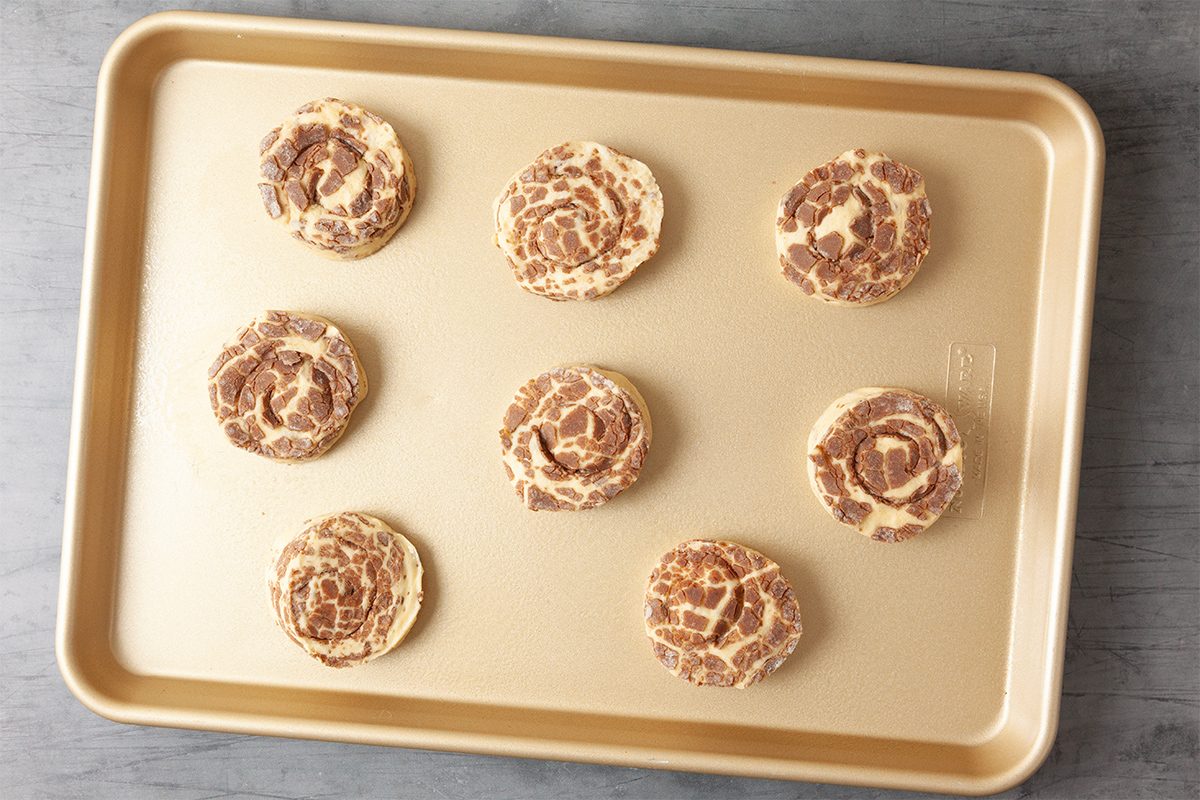 A baking tray with eight uncooked cinnamon rolls evenly spaced on its surface, ready for the oven. The rolls display a swirled pattern with visible cinnamon filling. The tray is placed on a light gray surface.