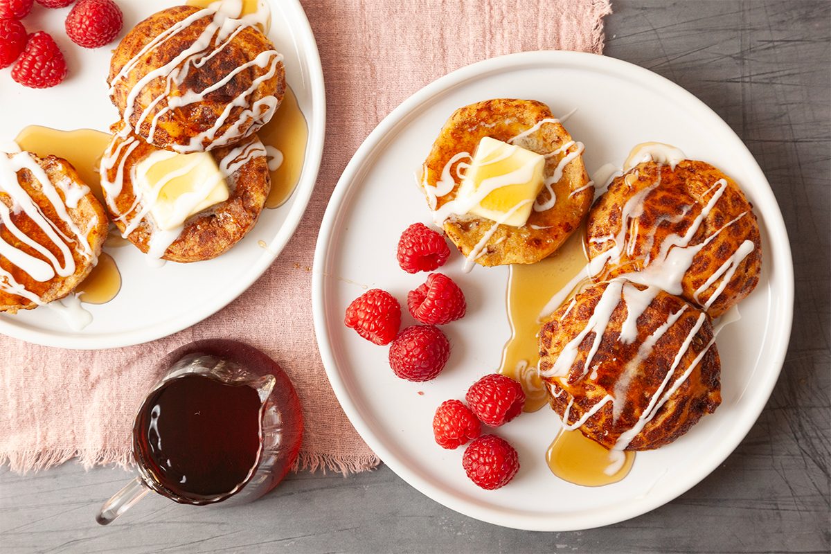 Two plates of pancakes topped with butter, drizzled with cream or icing, and syrup. Fresh raspberries are on the side. A small pitcher of syrup is nearby, all set on a pink cloth and gray tabletop.