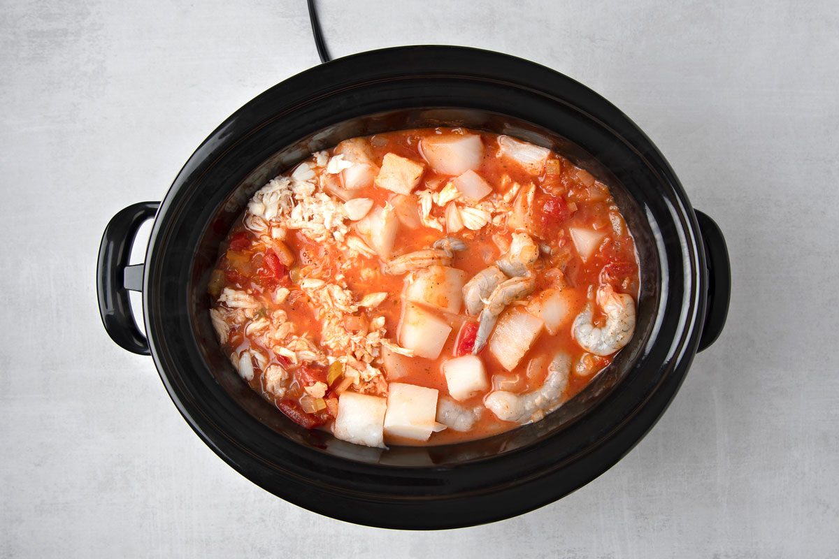 overhead shot of a black slow cooker filled with a red seafood stew; the stew contains diced potatoes, shrimp, and crab meat; the stew is set on a light gray countertop