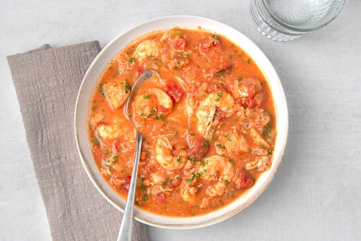 overhead shot of a bowl of seafood stew; the stew is a rich red color and contains large chunks of shrimp, fish, and vegetables; a spoon is in the bowl and a glass of water is next to the bowl; the bowl is on a light gray surface with a gray napkin to the left of the bowl