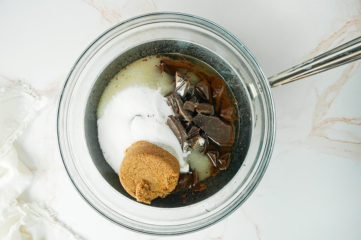 A glass bowl containing chunks of dark chocolate, granulated sugar, brown sugar, and a liquid ingredient, possibly melted butter. A mixing utensil is visible at the edge. The bowl is placed on a white marble surface.