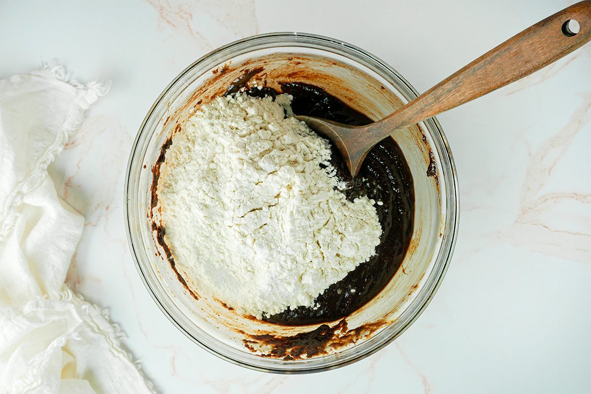 A glass bowl containing a mixture of flour and a dark liquid, likely batter, is being stirred with a wooden spoon. The bowl is placed on a light-colored surface, with a white cloth partially visible to the side.