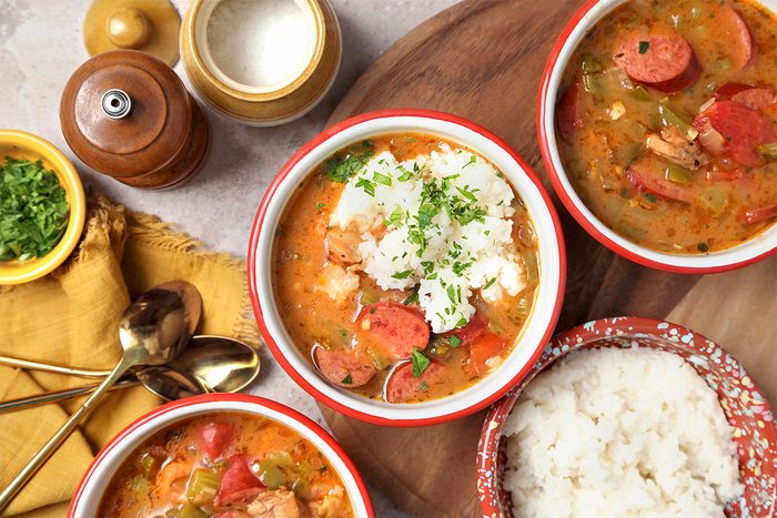 Three bowls of soup with sliced sausage, vegetables, and rice are garnished with parsley. A separate bowl of rice, a salt shaker, pepper grinder, and small dish of chopped herbs are nearby on a wooden surface.