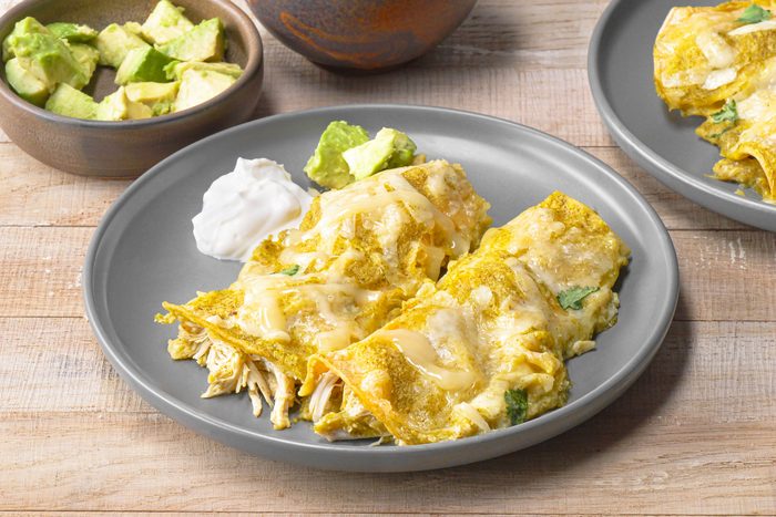 closeup of Enchiladas Verdes; two enchiladas are on a gray plate with a dollop of sour cream and avocado slices, the enchiladas are drizzled with a green sauce and cheese; there is a small bowl of avocado slices in the background; another bowl of what looks like sour cream is also in the background; both dishes are placed on a wooden table
