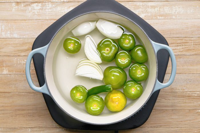 overhead shot of a light blue pot with a black handle on a black burner; inside the pot is water with green tomatoes, white onion slices, and a green jalapeno pepper; the pot sits on a wooden surface with a light wood grain pattern