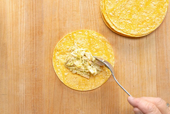 overhead shot of a person preparing a dish on a wooden cutting board; there is a large tortilla placed on the board and a hand is using a spoon to place a generous portion of shredded chicken and green sauce on the tortilla, another pile of smaller tortillas can be seen on the upper right corner of the board
