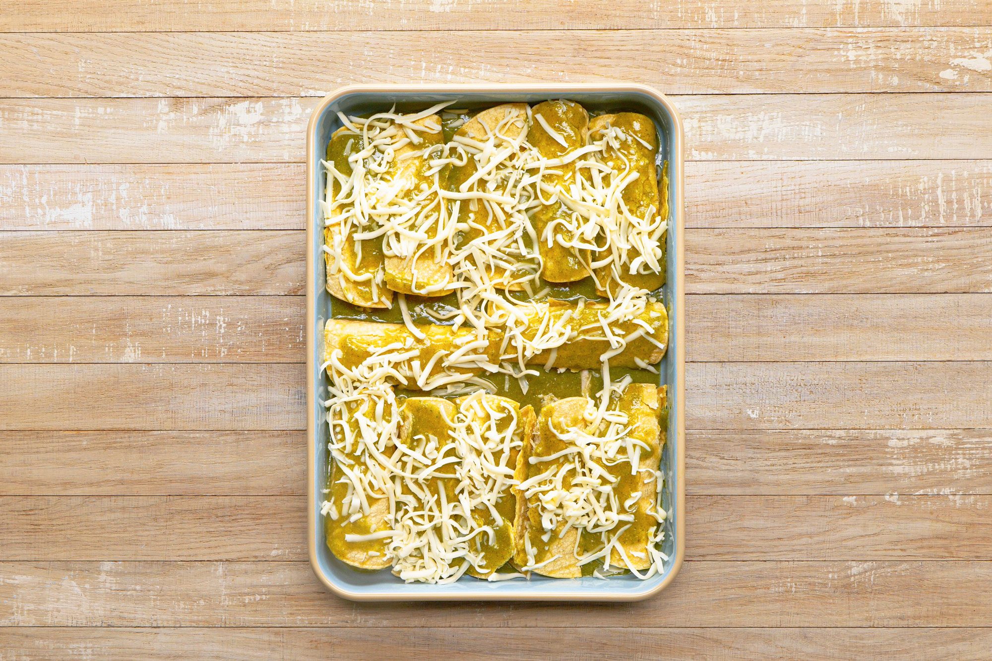 overhead shot of a baking dish filled with enchiladas, ready for baking; the enchiladas are topped with a green sauce and shredded cheese; the baking dish is sitting on a wooden surface