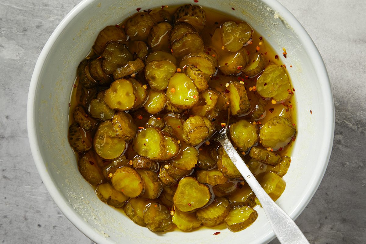 A white bowl filled with sliced pickles immersed in a spicy-looking liquid, with a spoon resting inside. The pickles are evenly distributed, and the liquid has visible seasoning specks. The background is a textured gray surface.