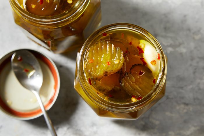 Top view of a jar filled with pickle slices, chili flakes, and garlic cloves. Nearby, there's a small bowl with a spoon resting inside. The scene is set against a gray textured background.