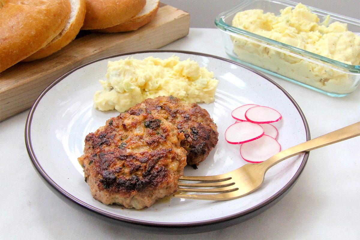 A plate with two browned meat patties, a side of mashed potatoes, and sliced radishes. A gold fork rests on the plate. In the background, a glass dish with mashed potatoes and a wooden board with bread rolls are visible.