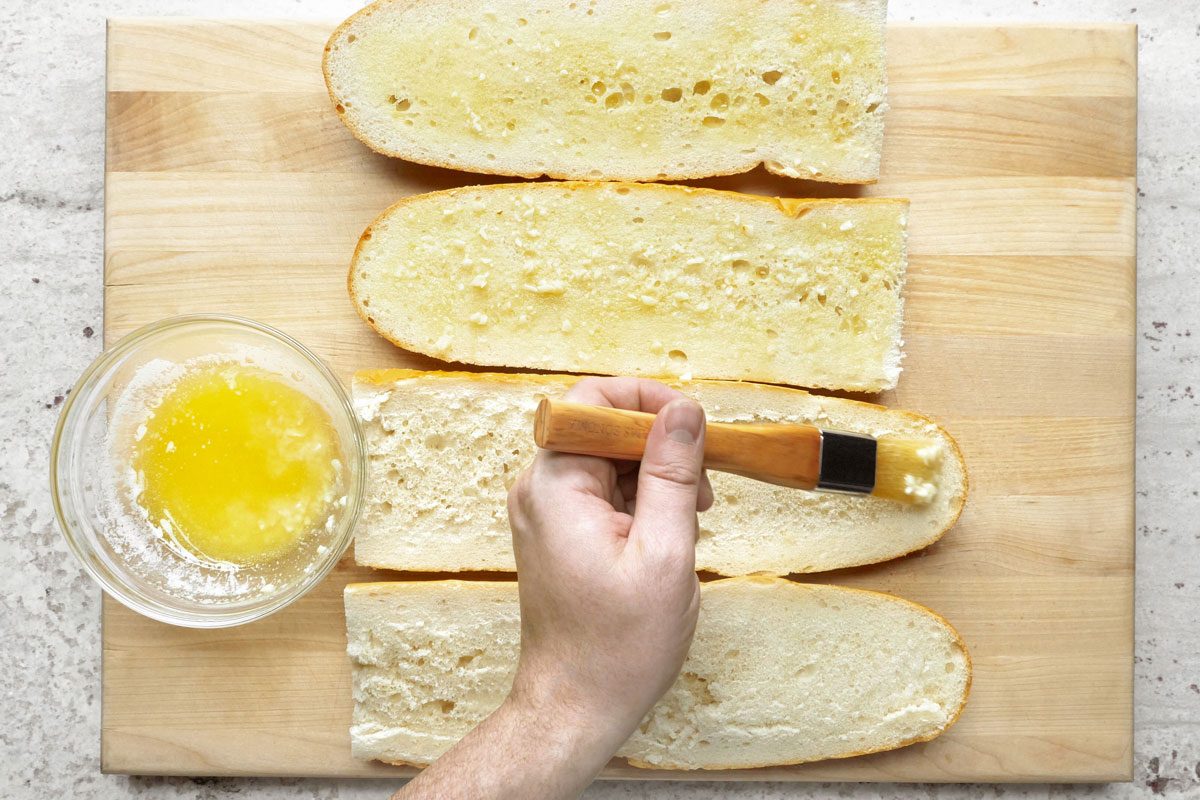 overhead shot of a wooden cutting board with four pieces of bread on it; a hand is holding a pastry brush and is applying melted butter to the bread; the bread is spread with garlic butter; the bread is on a wooden cutting board; a glass bowl with melted butter is in the lower left corner; a white speckled countertop is to the right of the cutting board