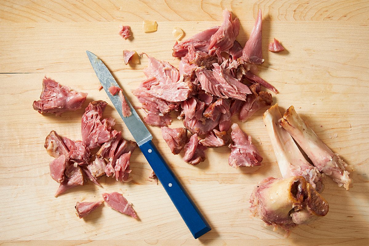 Chopped pieces of cooked ham on a wooden cutting board with a blue-handled knife beside them.