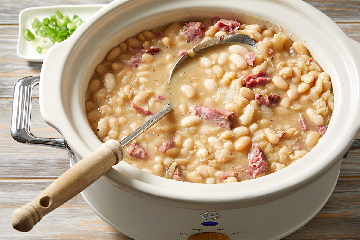 A slow cooker filled with creamy white beans and chunks of ham. A ladle rests inside, and a small dish with chopped green onions is seen in the background on a wooden surface.