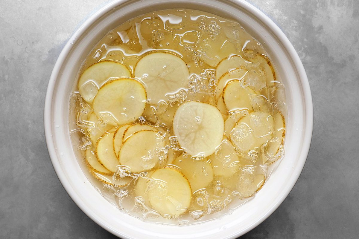Top view of thinly sliced potatoes in a white bowl filled with ice water, on a gray surface. The slices are arranged in overlapping layers, and ice cubes are visible around them.