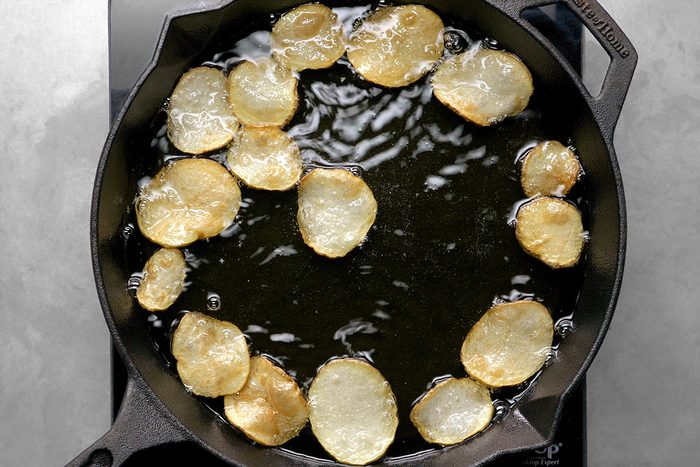 Potato slices frying in hot oil in a black cast iron skillet. The potato slices are golden brown and sizzling, surrounded by bubbles from the cooking oil. The skillet is on a gray stovetop surface.