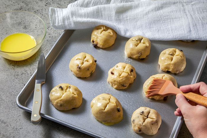 3/4th shot of a baking tray with eight round, golden-brown hot cross buns on it, arranged in two rows of four, a hand holding a pastry brush is seen brushing melted butter over the top of one bun; there is a white cloth in the background, a bowl of melted butter and a knife is seen on the left side of the baking tray; the baking tray is on a grey surface