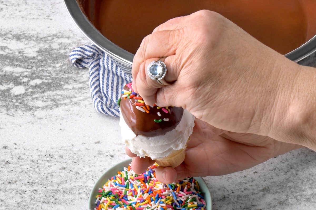 overhead shot of a person is holding a chocolate-covered ice cream cone over a bowl of sprinkles; the ice cream cone is covered in chocolate and sprinkles