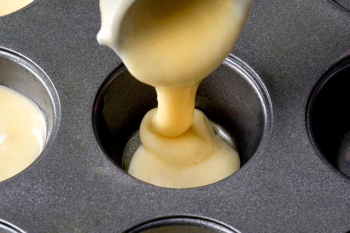 a close-up image of a muffin tin being filled with batter, the batter is being poured from a pitcher and it is flowing into a muffin tin , the batter is a light yellow color; the muffin tin is dark gray in color and has a non-stick coating