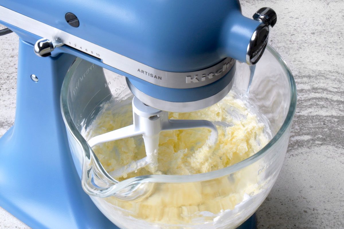 3/4th shot of a blue stand mixer with a glass bowl attached is shown in the image; the mixer is on a gray countertop; the glass bowl contains a mixture of ingredients