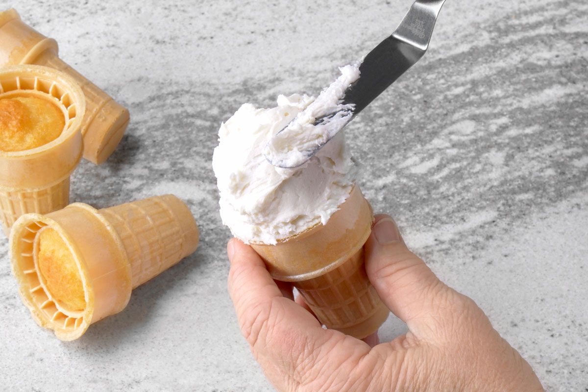 overhead shot of a hand holding an ice cream cone with frosting on top of it; the frosting is being spread with a metal spatula; there are two other ice cream cones laying flat on the counter to the left of the hand; the background is a light grey countertop